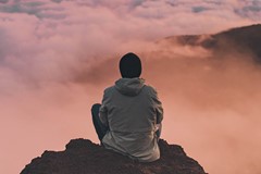 person in jacket and hat sitting on mountaintop above clouds