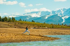 woman in warrior ii yoga pose next to river with mountains and trees in background