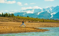 woman in warrior ii yoga pose next to river with mountains and trees in background