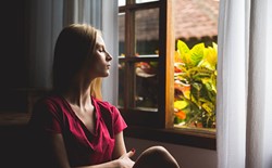 woman with eyes closed sitting next to open window with plants outside